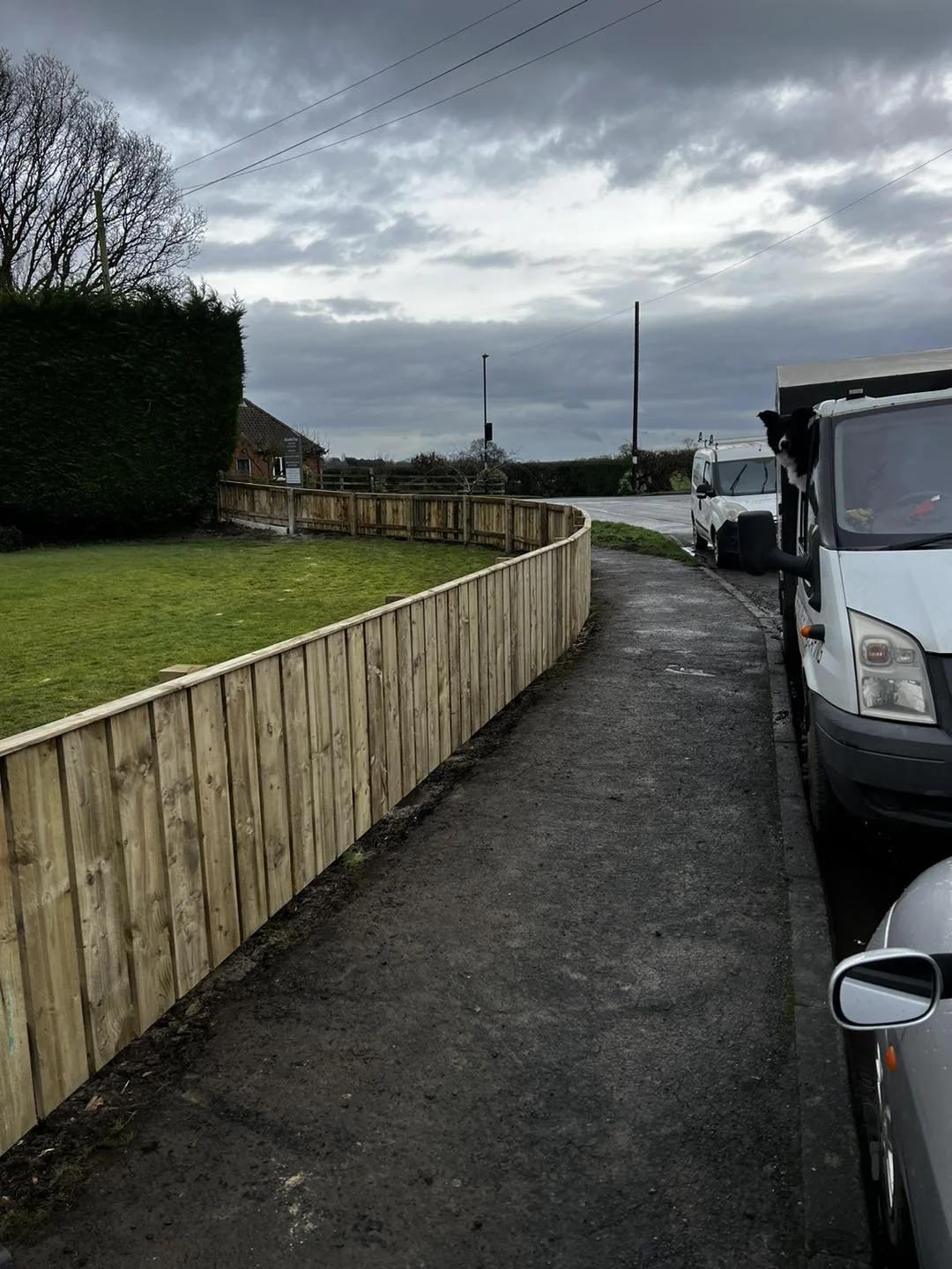 Curved featheredge fence along a roadside verge, with the team's dog watching from the van